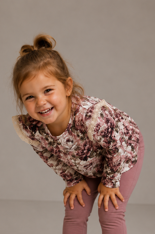 A young girl with a high bun is crouching down in a studio setting, wearing a floral long-sleeve top and dusty rose flared leggings.