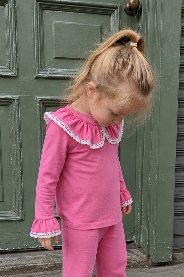 A young girl with blonde hair in a ponytail stands on a sidewalk in front of a green paneled door and a gray metal shutter. She is wearing a bright pink long-sleeve top and matching pants with white ruffled trim on the collar and cuffs, along with white sneakers.