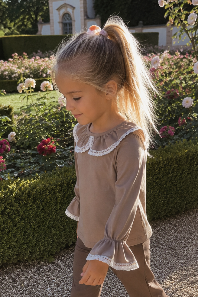 A young girl with blonde hair in a ponytail walks along a gravel path in a formal garden, wearing a matching brown two-piece outfit with white lace trim on the top and ruffled hems on the pants.