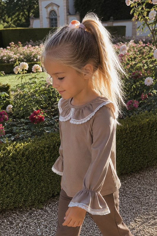 A young girl with blonde hair in a ponytail walks along a gravel path in a formal garden, wearing a matching brown two-piece outfit with white lace trim on the top and ruffled hems on the pants.
