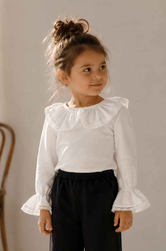 A young girl stands in a bright, sunlit room wearing a white long-sleeve blouse with a ruffled collar and cuffs, paired with black wide-leg pants featuring ruffled hems, and black flats.