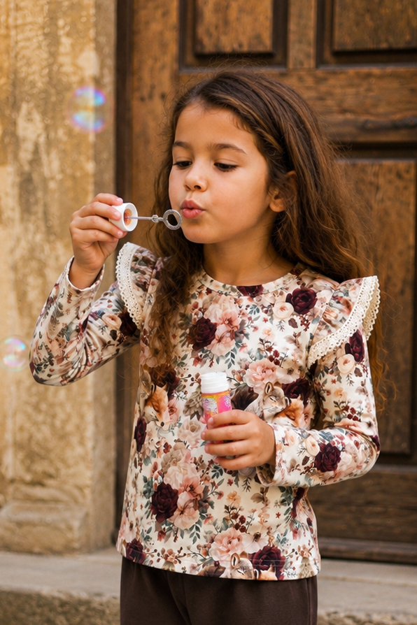 A young girl stands on stone steps in front of a rustic stone wall and wooden door, wearing a long-sleeve floral top with ruffled shoulders and dark brown flared pants.