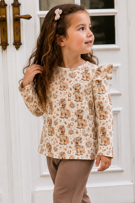 A young girl with long wavy brown hair wearing a beige and brown teddy bear patterned long-sleeve top with ruffle details on the shoulders, paired with brown ruffled pants, standing in front of white paneled doors.