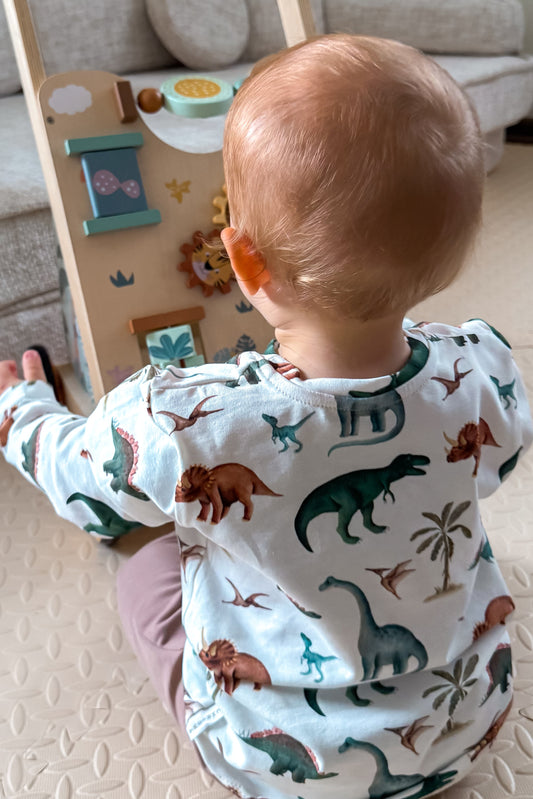 Baby in a dinosaur-themed outfit standing in a room with a couch and wall decorations.