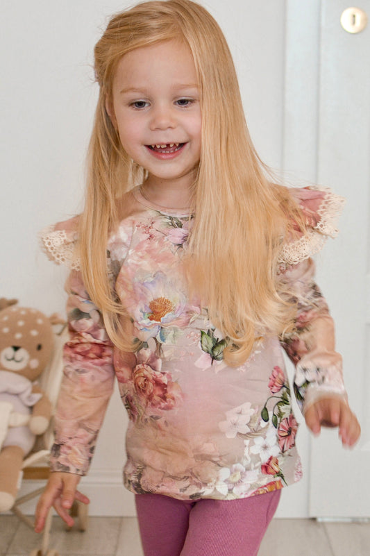 Young girl in a floral top and pink pants standing indoors with a teddy bear in the background.