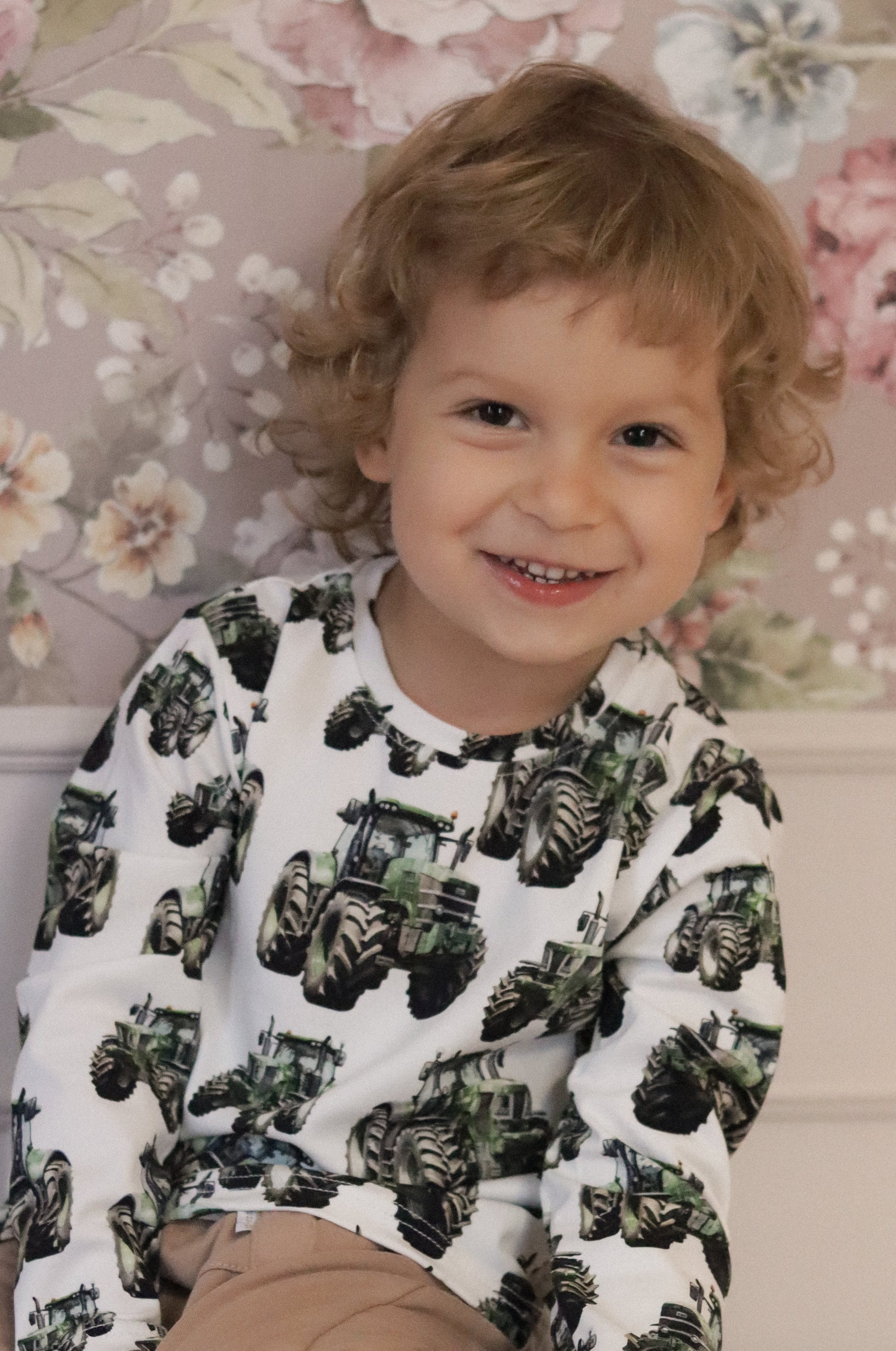 Child wearing a long-sleeve shirt with tractor prints against a floral wallpaper background