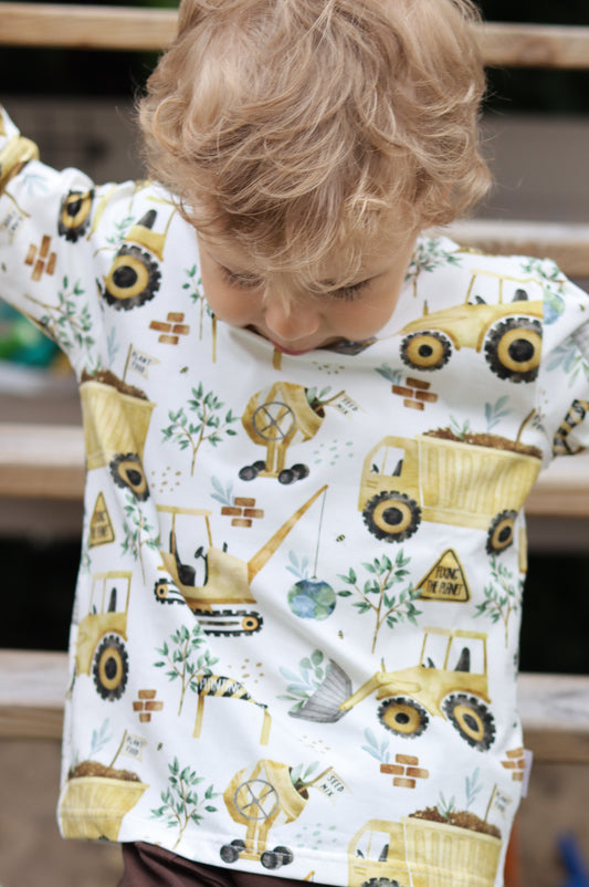 Child wearing a onesie with tractor pattern, standing in front of a wooden fence.