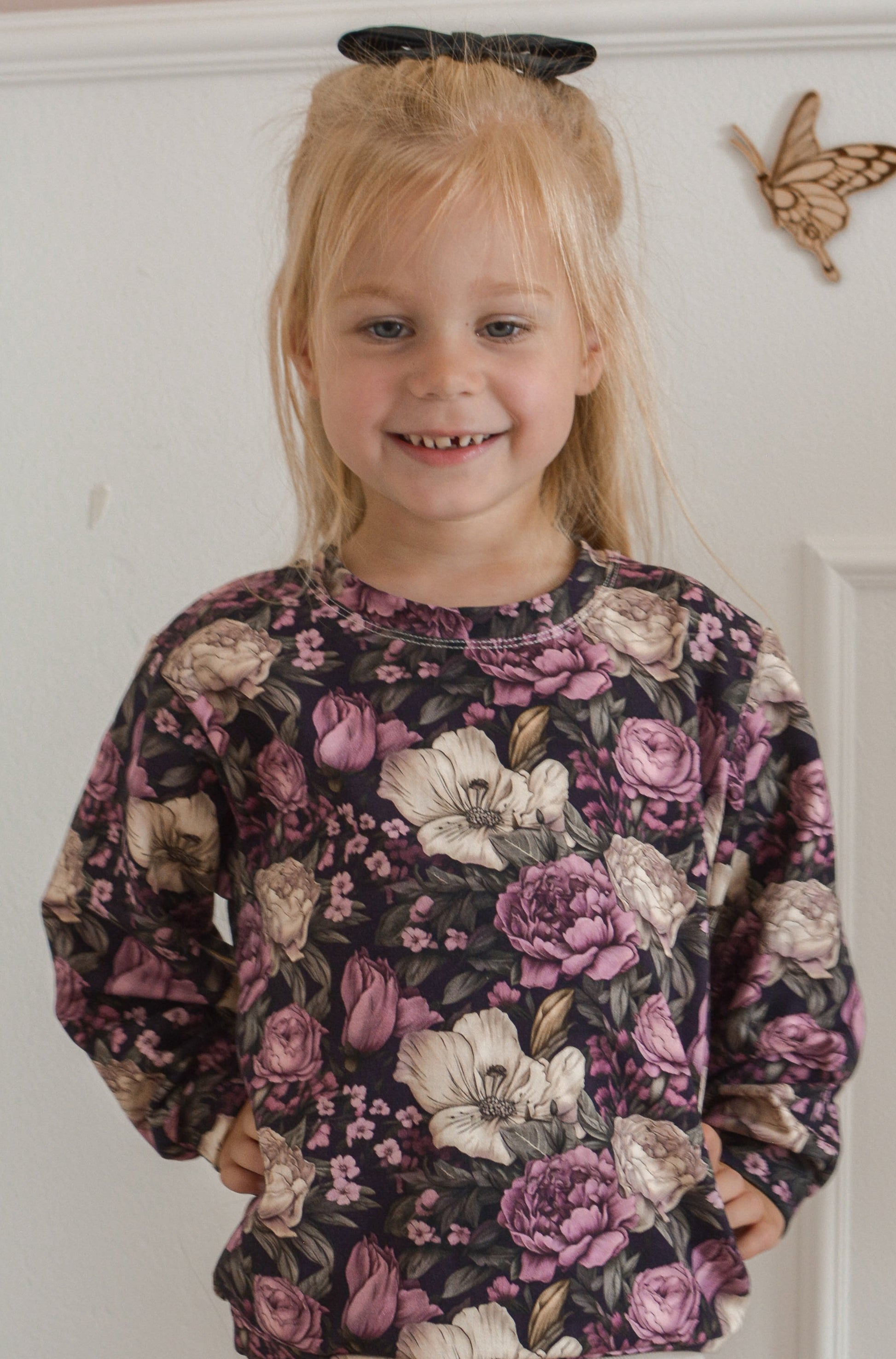 Young girl wearing a floral sweater and white pants standing in a room with a white wall and beige chair.
