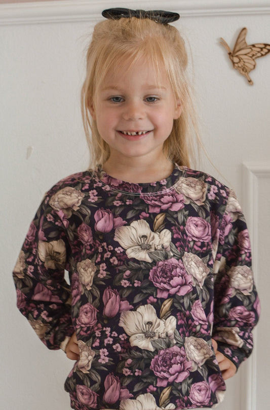 Young girl wearing a floral sweater and white pants standing in a room with a white wall and beige chair.