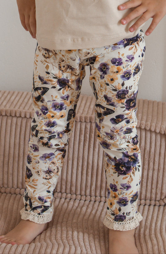 Young girl in a beige top and floral pants standing in a room with a white wall and pink cushion.
