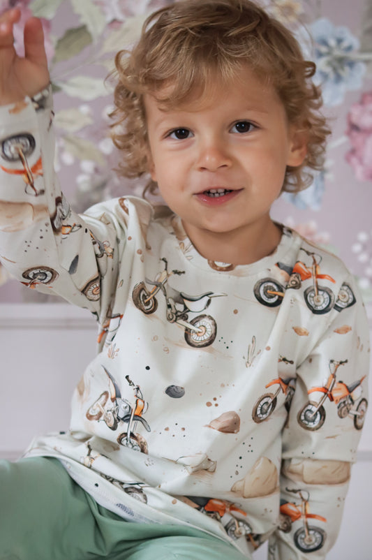 Child wearing a long-sleeve shirt with bicycle print and light green pants against a floral background
