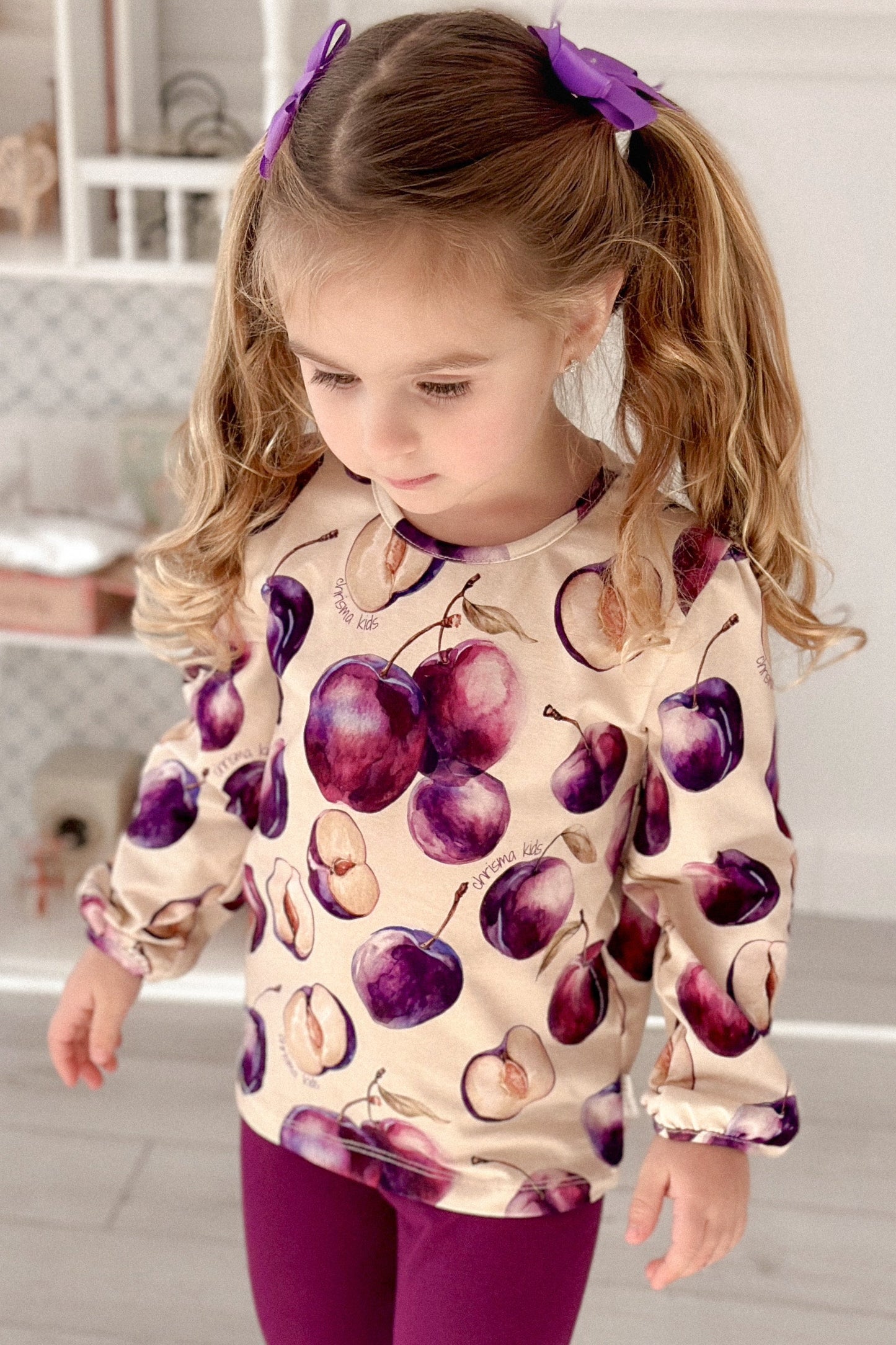 Child wearing a sweater with plum pattern and purple pants in a room.
