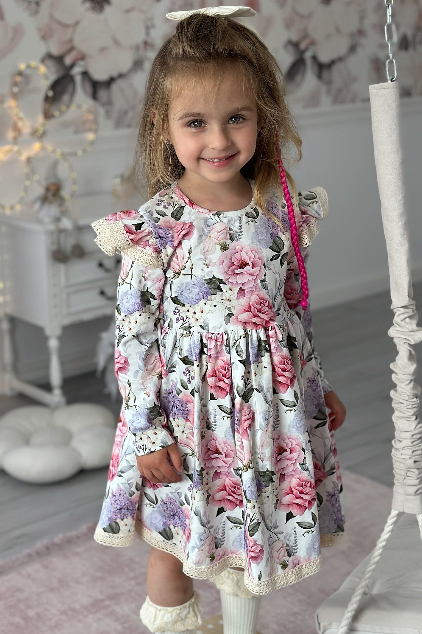 Young girl in a floral dress standing in a decorated room with a swing and fairy lights.